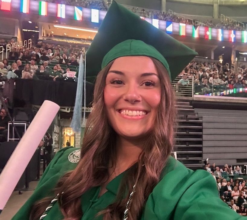 Ellie in a green cap and gown at her Michigan State graduation, holding her diploma.